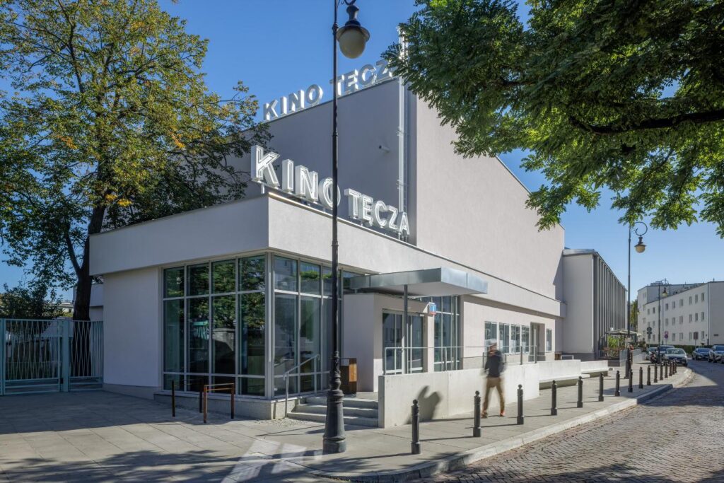 Budynek kina z napisem „KINO TĘCZA” nad wejściem, otoczony drzewami przy ulicy.