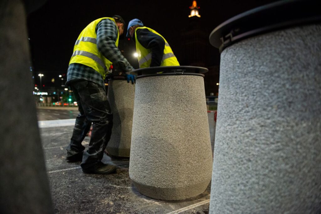 Dwóch pracowników w kamizelkach odblaskowych ustawia betonowe kosze na nocnym chodniku.