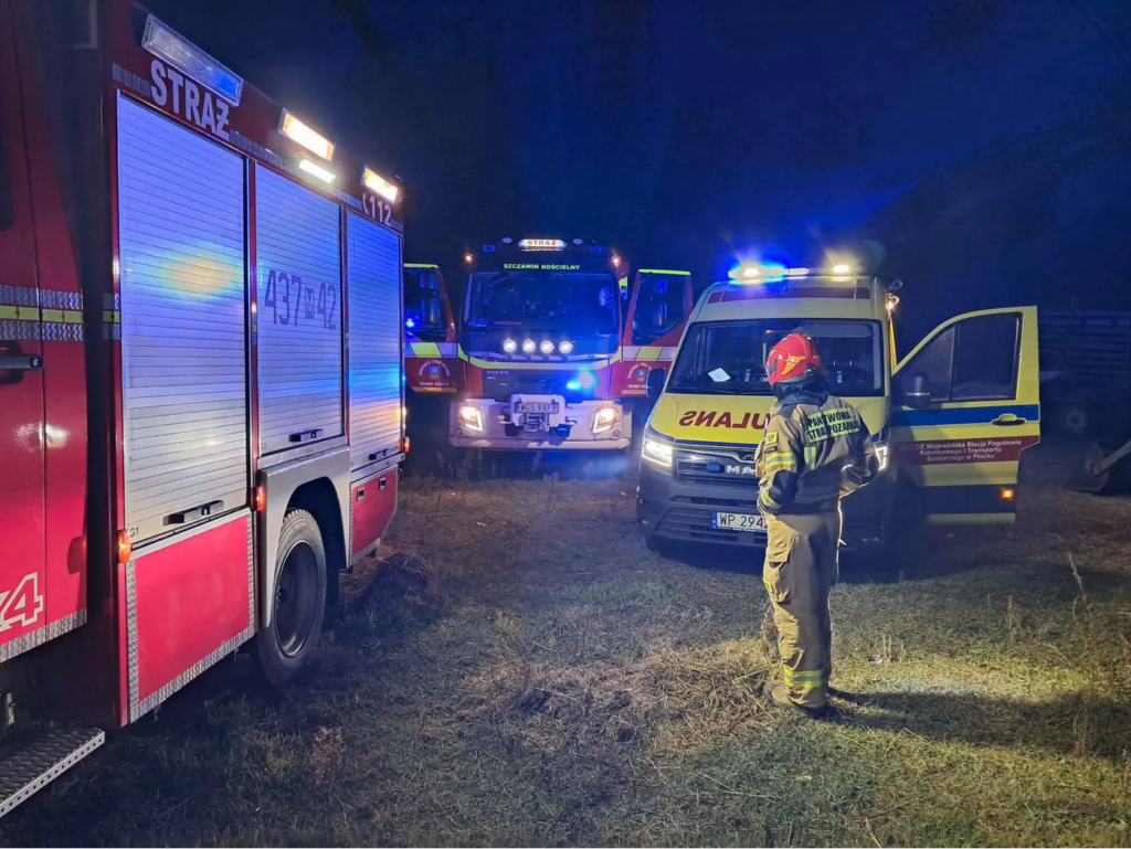 Strażak stoi przy otwartych drzwiach karetki, obok wozów strażackich na oświetlonym polu nocą.