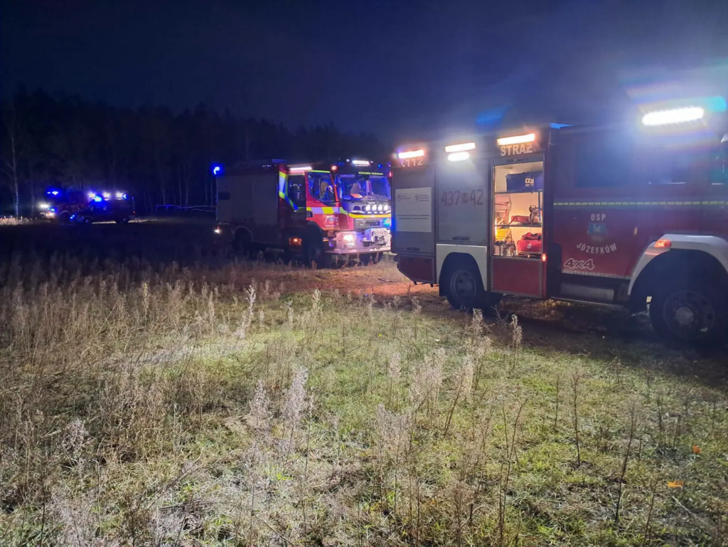 Wozy strażackie z włączonymi sygnałami świetlnymi stoją nocą na polu przy zaroślach.