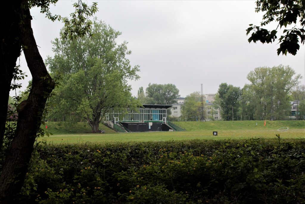 Zielony teren sportowy z trawiastym boiskiem, bramką i niskim budynkiem pośród drzew.