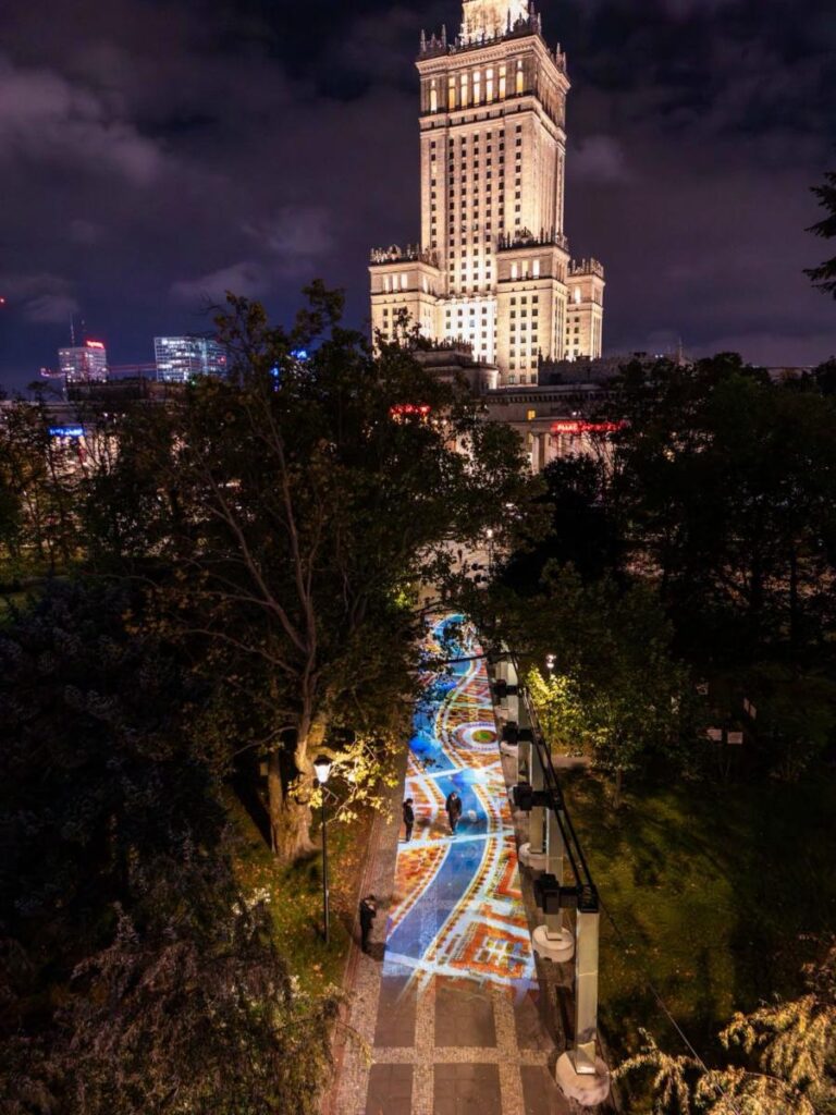 Kolorowa iluminacja świetlna na ścieżce między drzewami, w tle oświetlony wieżowiec nocą.