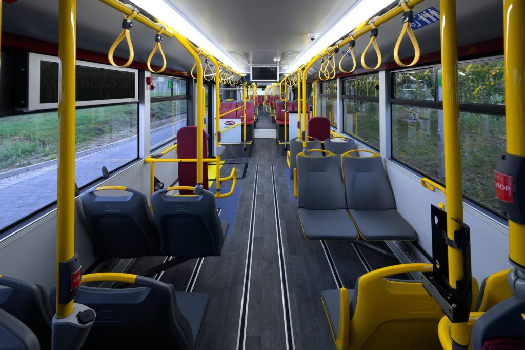 Puste wnętrze autobusu z żółtymi poręczami, szarymi siedzeniami i dużymi oknami, widok wzdłuż przejścia.