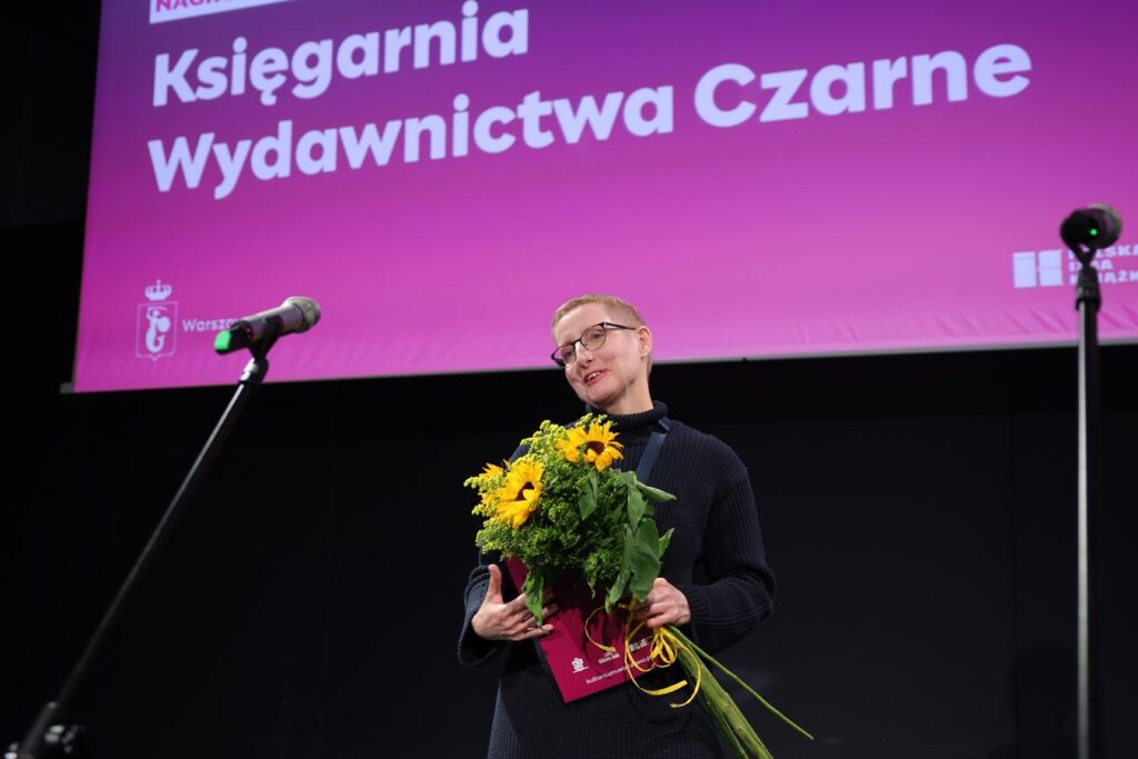 Osoba w okularach trzyma bukiet słoneczników na scenie, na tle napisu „Księgarnia Wydawnictwa Czarne”.