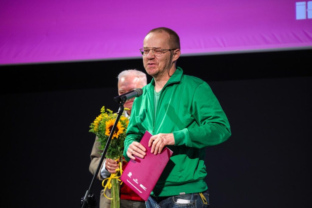 Mężczyzna w zielonej bluzie przemawia przy mikrofonie na scenie, trzymając różową teczkę.