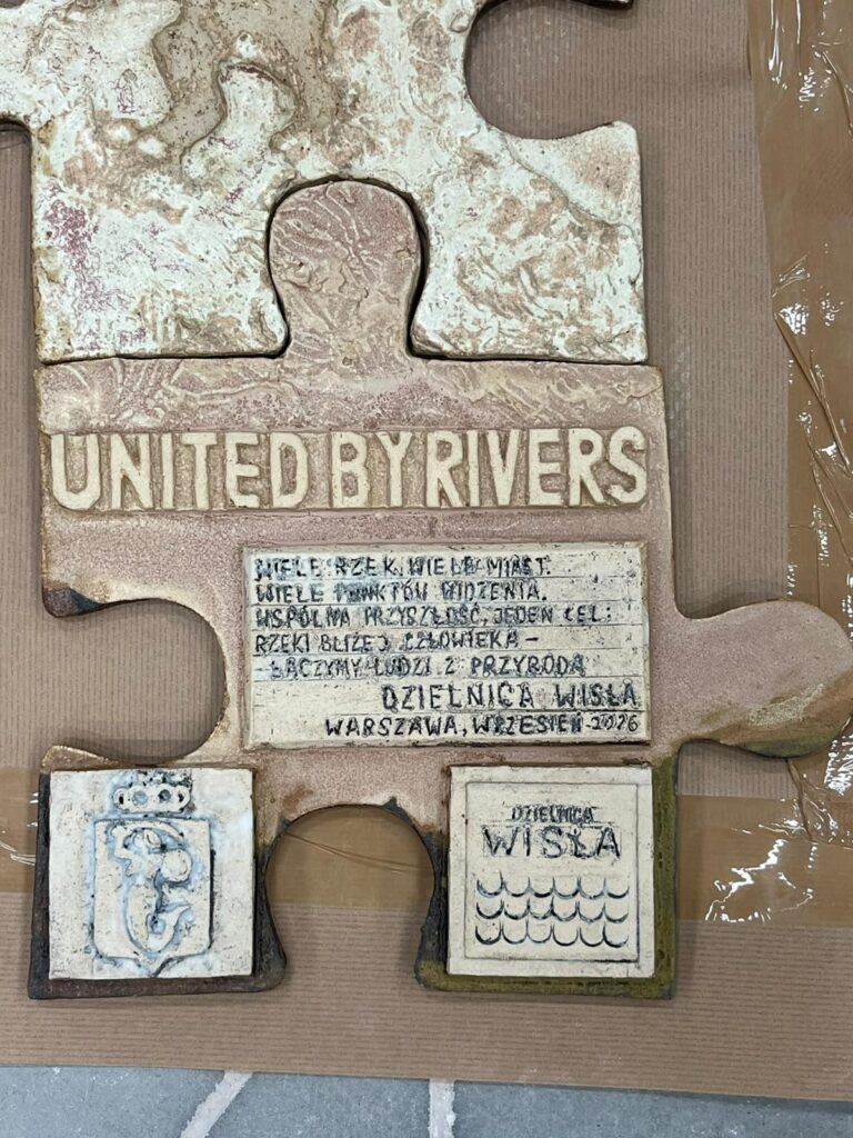 Ceramiczny element mozaiki w kształcie puzzla z napisem „UNITED BY RIVERS” i tabliczkami na kartonowym podłożu.