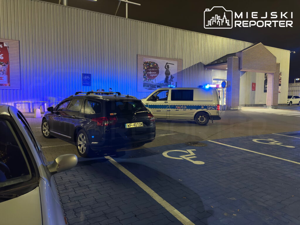 Radiowóz z włączonymi niebieskimi światłami stoi obok samochodów na parkingu przed oświetlonym budynkiem.