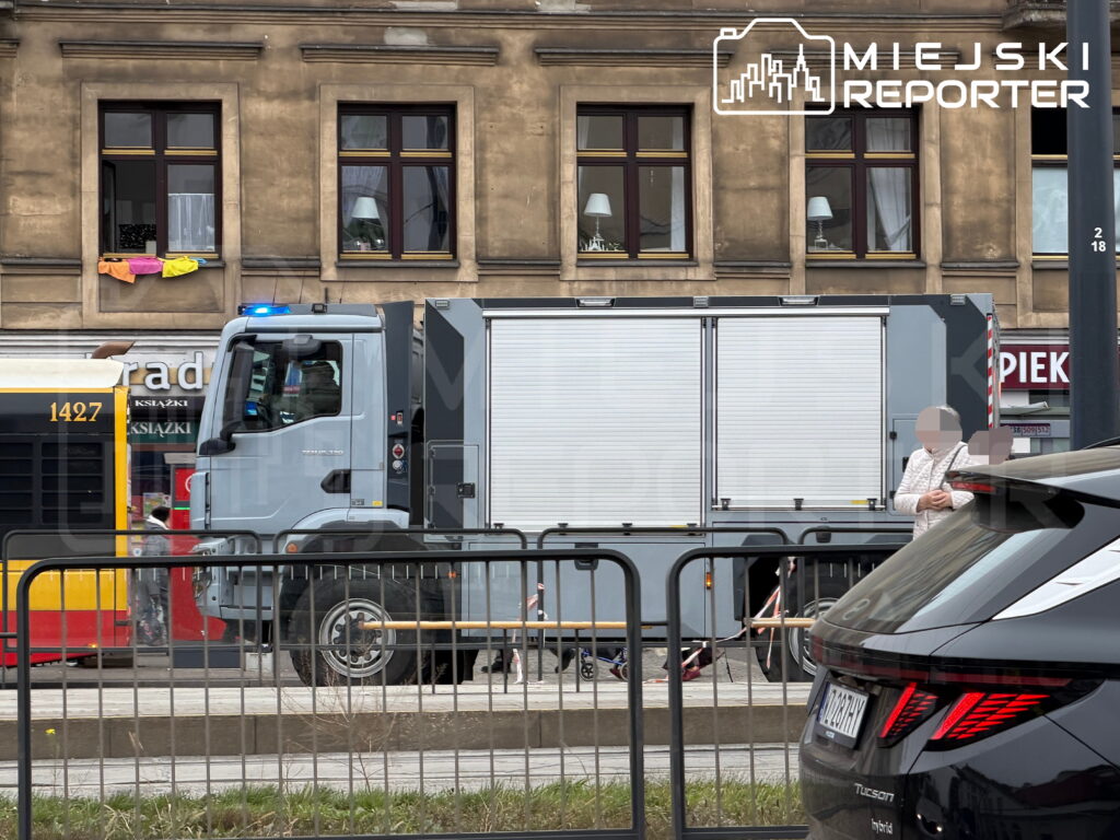 Szary samochód służb ratunkowych z włączonym niebieskim światłem na ulicy, przed kamienicą i za barierkami