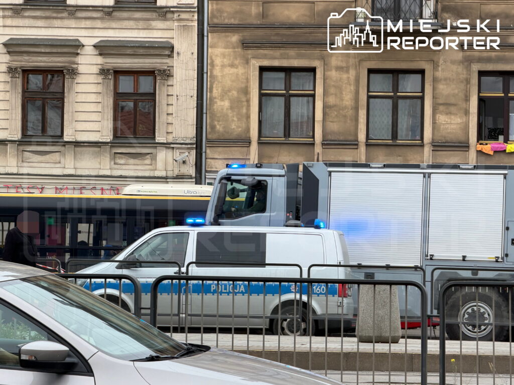 Radiowóz policji z włączonymi niebieskimi światłami i ciężarówka za barierkami na ulicy wśród kamienic.
