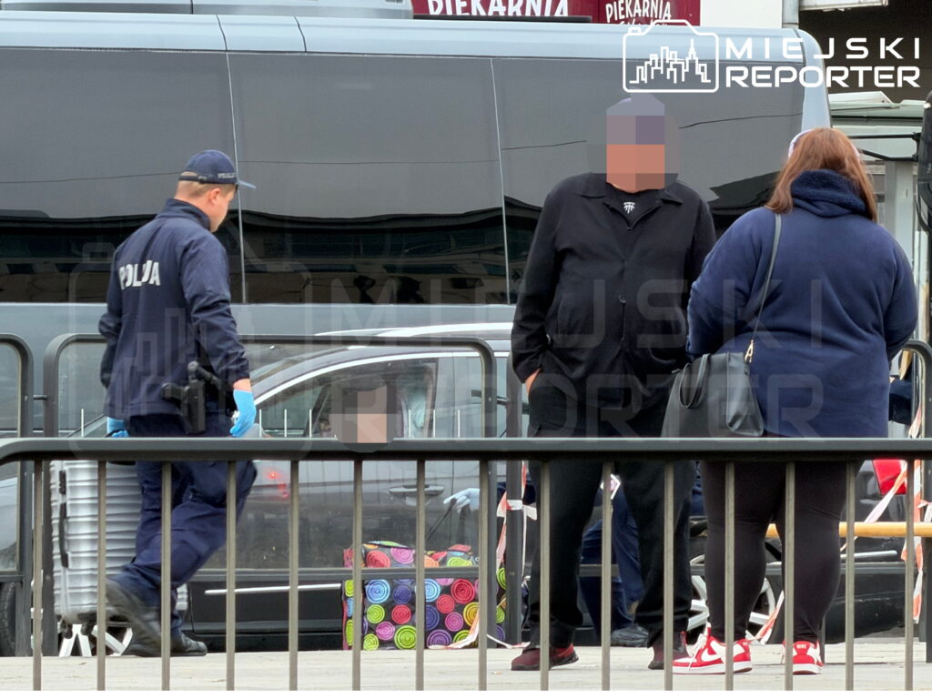 Policjant w rękawiczkach przechodzi obok grupy osób przy barierkach przed zaparkowanym tramwajem.