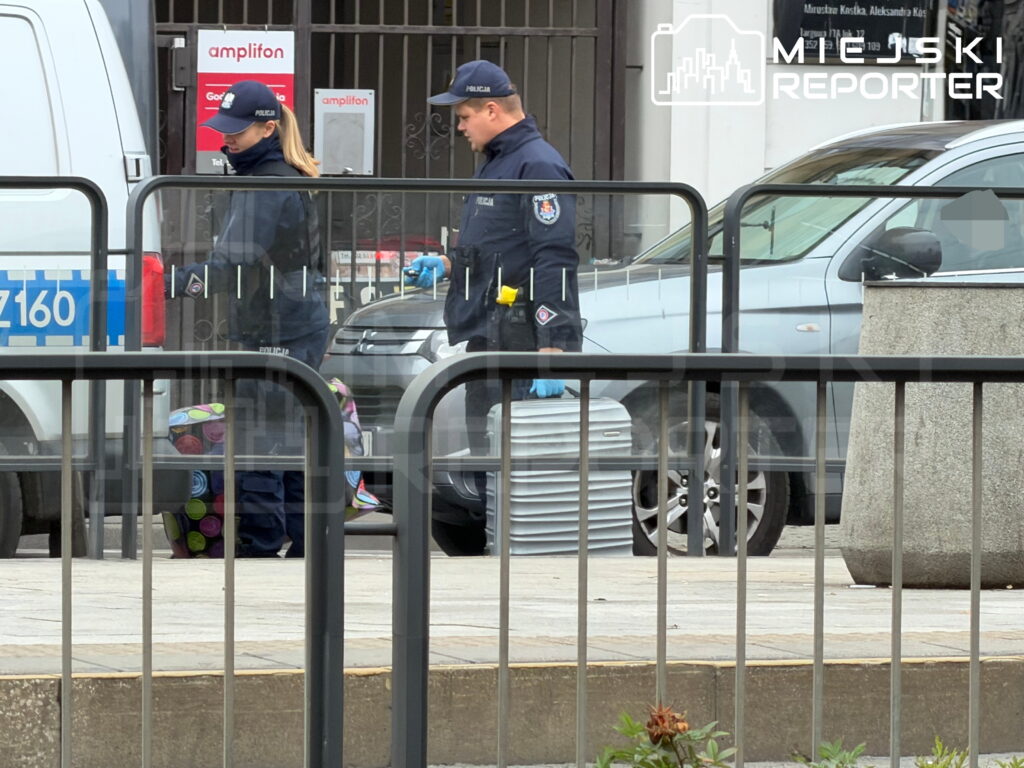 Dwoje policjantów w rękawiczkach ogląda przedmiot przy zaparkowanych samochodach, za barierkami ulicznymi.