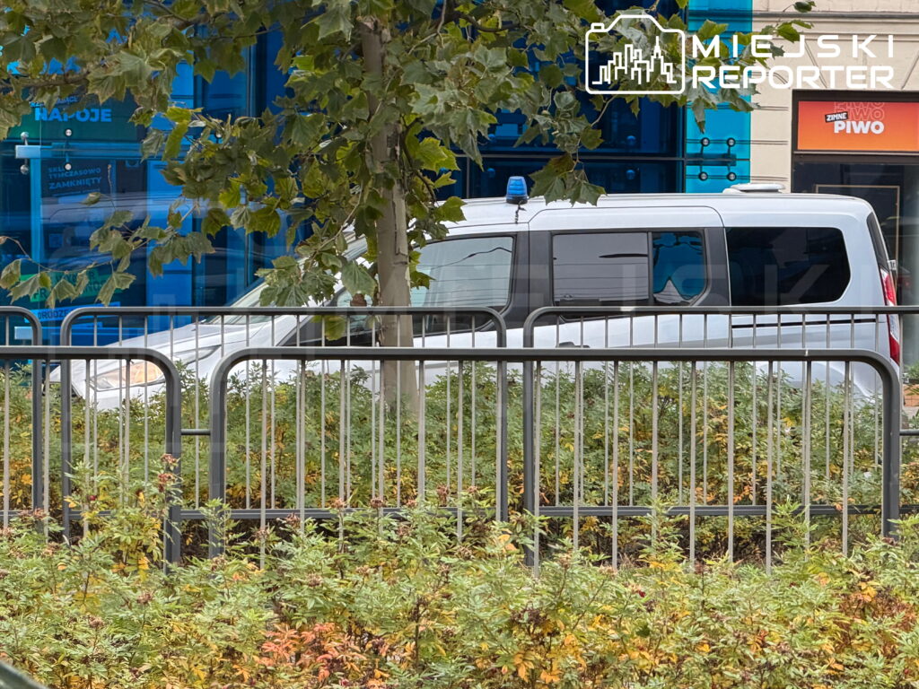 Biały policyjny van z niebieskim sygnałem świetlnym stoi za metalowymi barierkami przed przeszklonym budynkiem.