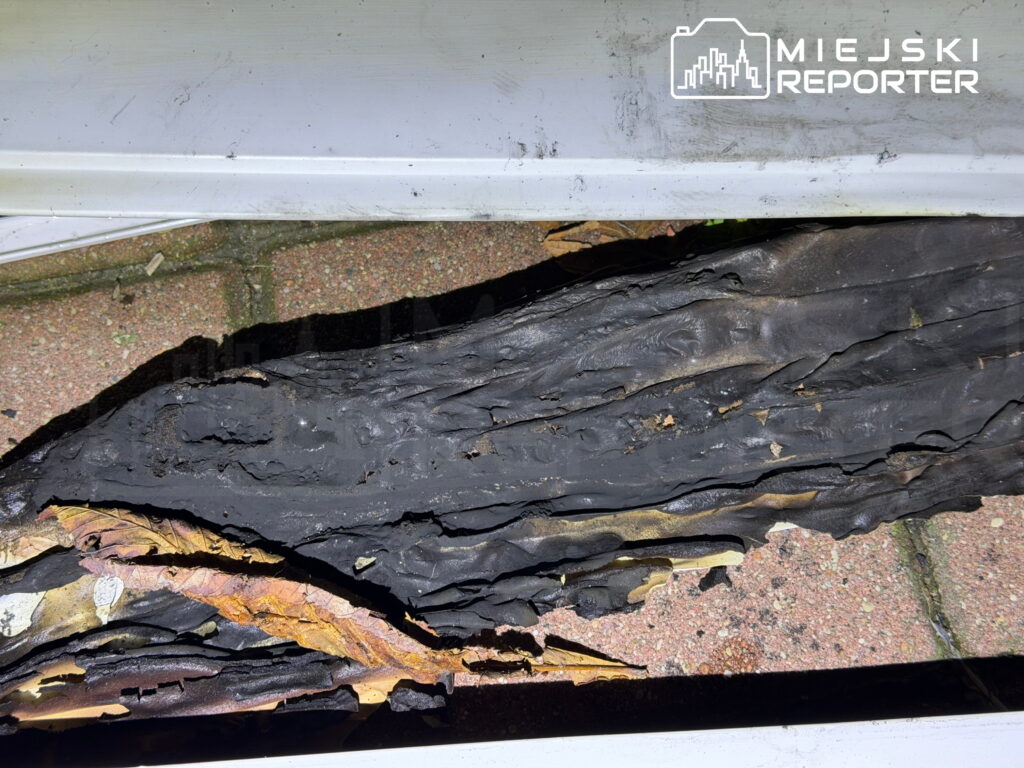 Spalone i zwęglone fragmenty materiału leżą na kamiennej posadzce przy metalowym progu.