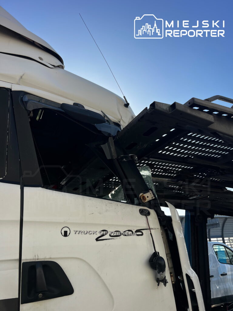 Uszkodzona kabina białej ciężarówki pod metalową rampą transportową, z pękniętą szybą i zgniecionymi elementami nadwozia.