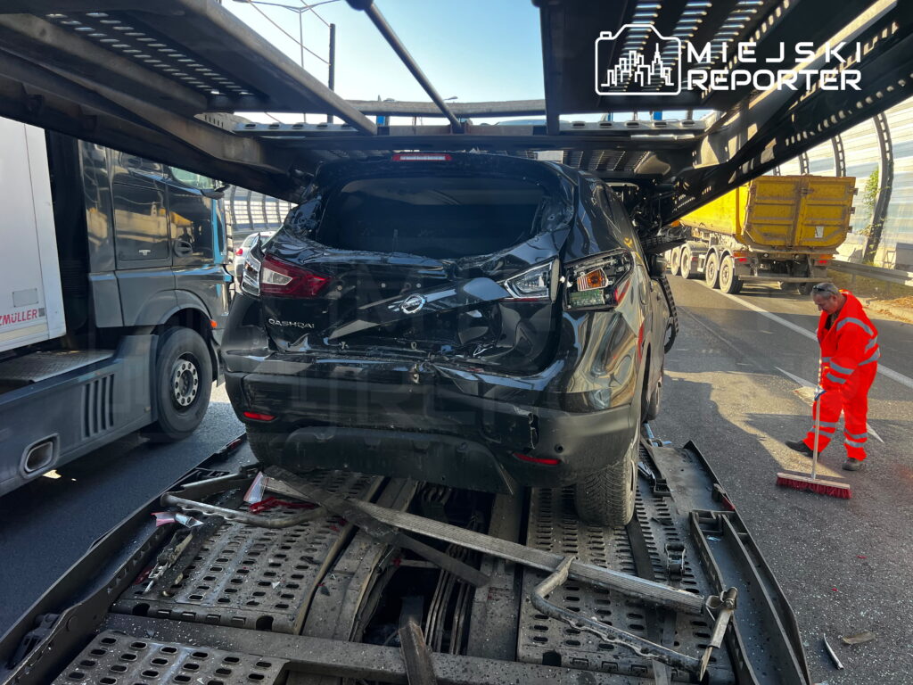 Uszkodzony czarny SUV na platformie lawety, obok pracownik służb porządkowych zamiatający jezdnię.