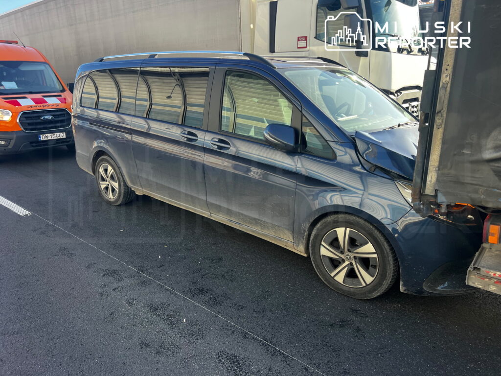 Granatowy minibus z uszkodzonym przodem stoi między ciężarówkami na jezdni.
