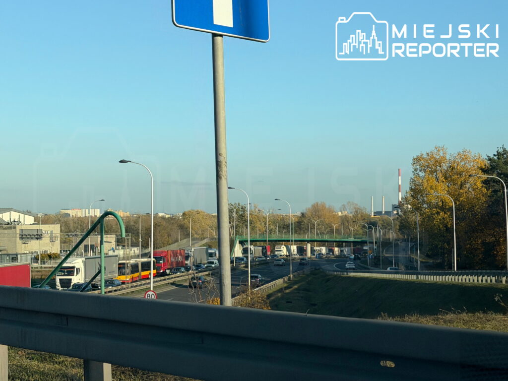 Korki na wielopasmowej drodze, gdzie samochody ciężarowe i osobowe jadą pod wiaduktem wśród jesiennych drzew.