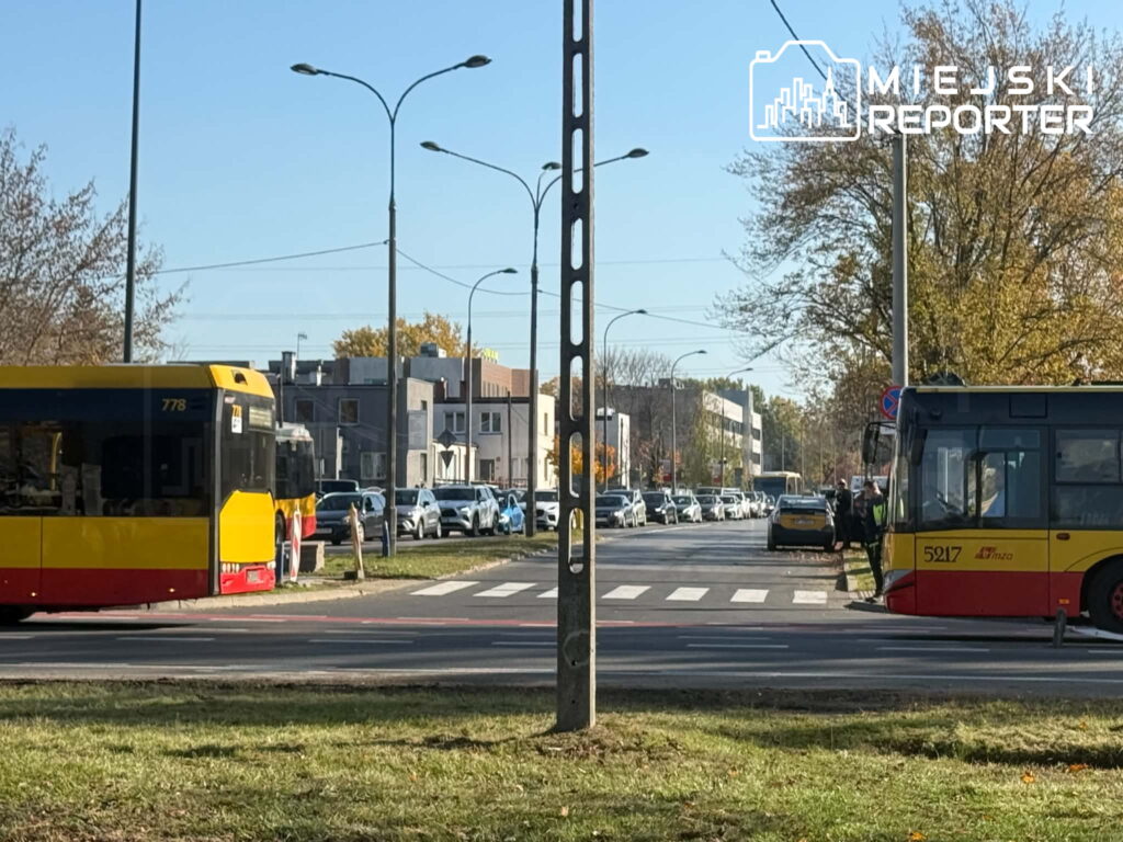 Dwa żółto-czerwone autobusy miejskie stoją przy przejściu dla pieszych, a samochody poruszają się ulicą w tle.