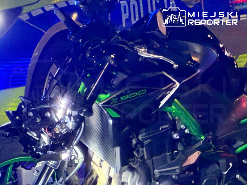 Uszkodzony zielono-czarny motocykl Kawasaki Z900 stoi nocą obok radiowozu policyjnego z włączonymi niebieskimi światłami.