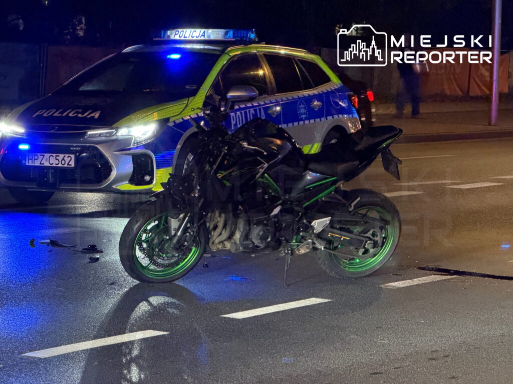 Uszkodzony motocykl stoi na jezdni przed radiowozem policji z włączonymi niebieskimi światłami.