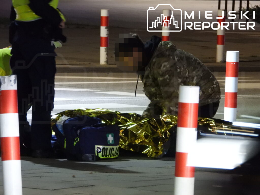 Ratownik w odzieży moro klęczy przy osobie okrytej złotą folią, obok stoją służby przy przejściu dla pieszych.