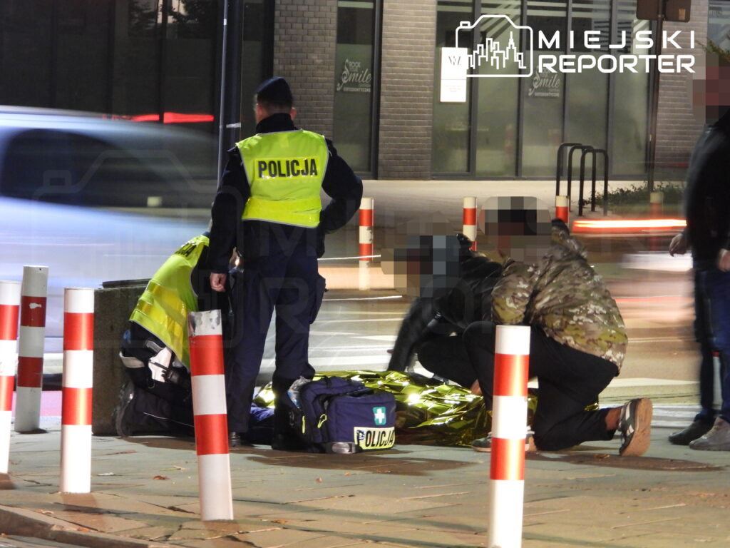 Policjanci w odblaskowych kamizelkach zabezpieczają miejsce zdarzenia na ulicy, przy leżącej osobie.