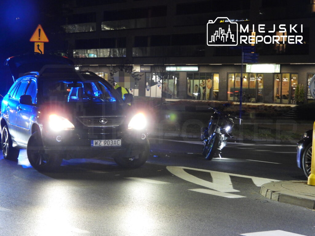 Ciemny SUV z włączonymi światłami stoi na skrzyżowaniu nocą, obok leży przewrócony motocykl.