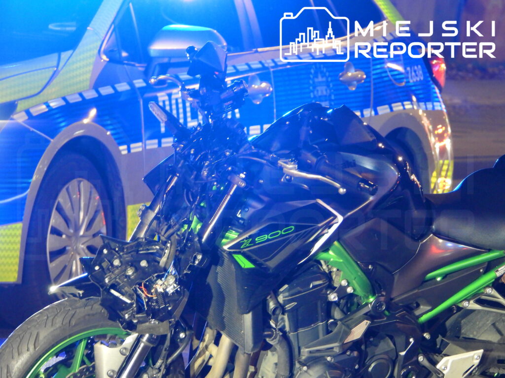 Uszkodzony czarno-zielony motocykl stoi przed oznakowanym radiowozem, oświetlony niebieskimi światłami.