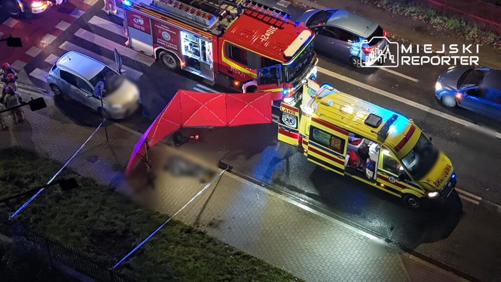 Straż pożarna i karetka stoją przy rozstawionym czerwonym parawanie na ulicy nocą.