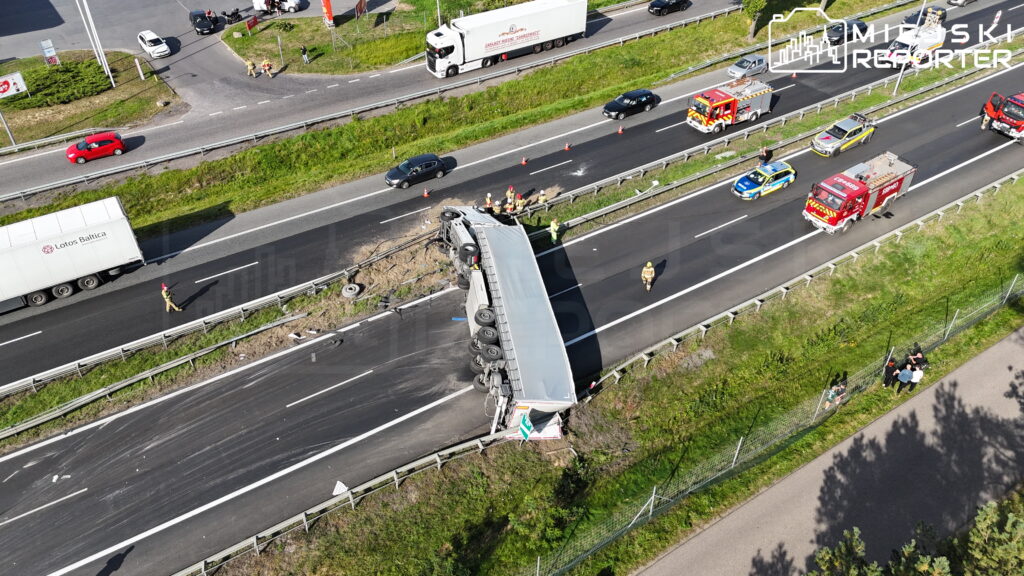 Przewrócona ciężarówka blokuje pasy autostrady, a obok pracują strażacy i policjanci.