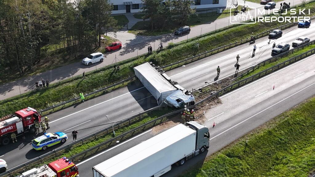 Ciężarówka przewrócona na barierach autostrady, obok pracują służby ratunkowe i stoją wozy strażackie.