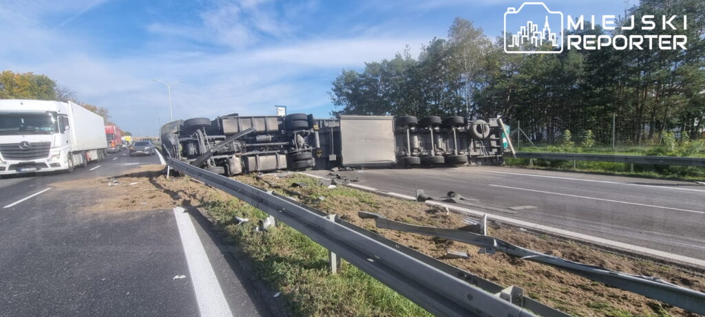 Przewrócona ciężarówka leży na boku przy barierze rozdzielającej jezdnie, obok jadą inne pojazdy.