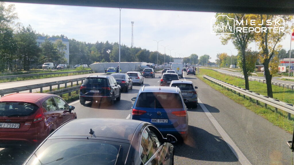 Kilkanaście samochodów stoi w korku na wielopasmowej drodze, wśród drzew i przydrożnych barier.