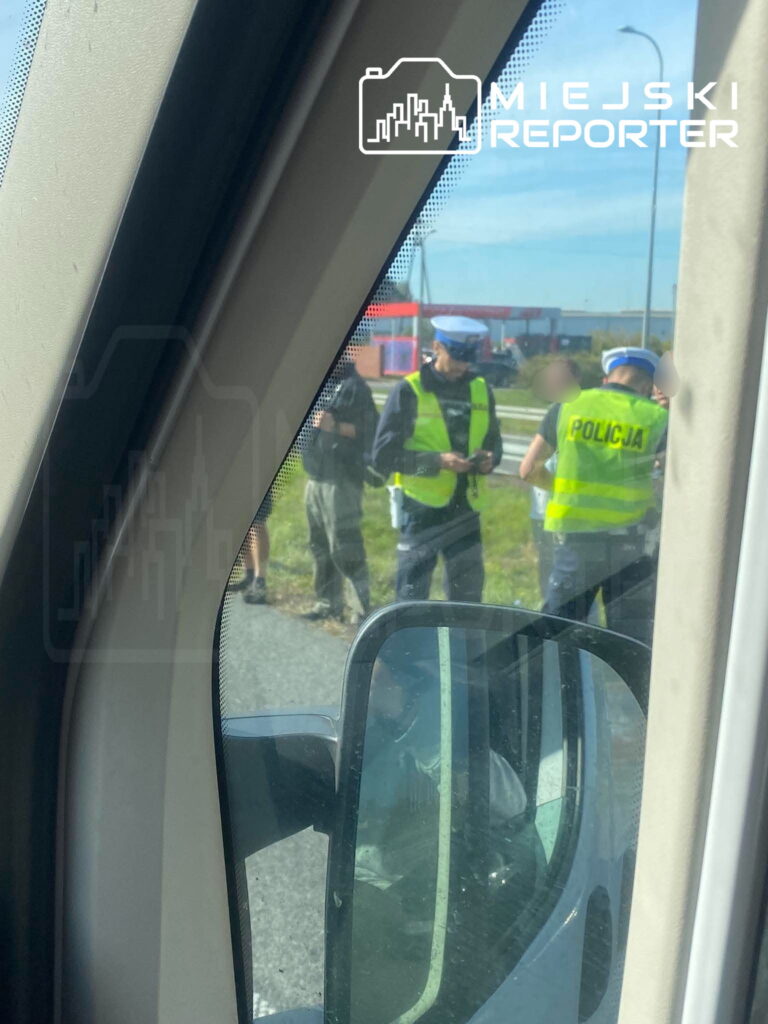 Policjanci w odblaskowych kamizelkach rozmawiają z osobami przy drodze, widziani przez okno samochodu.