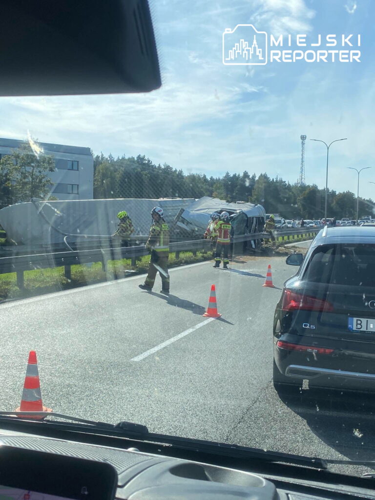 Strażacy zabezpieczają przewróconą ciężarówkę na drodze, gdzie ruch wyznaczają pachołki.