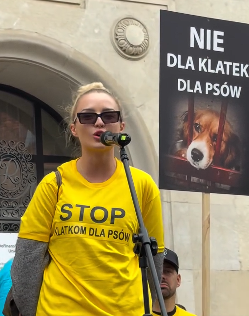 Kobieta w żółtej koszulce przemawia do mikrofonu; obok plakat z napisem „NIE DLA KLATEK DLA PSÓW”.