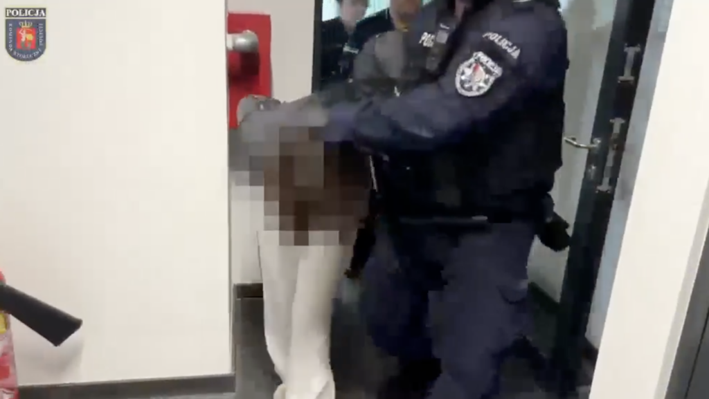 Policjanci prowadzą zamazaną osobę przez otwarte drzwi w korytarzu budynku.