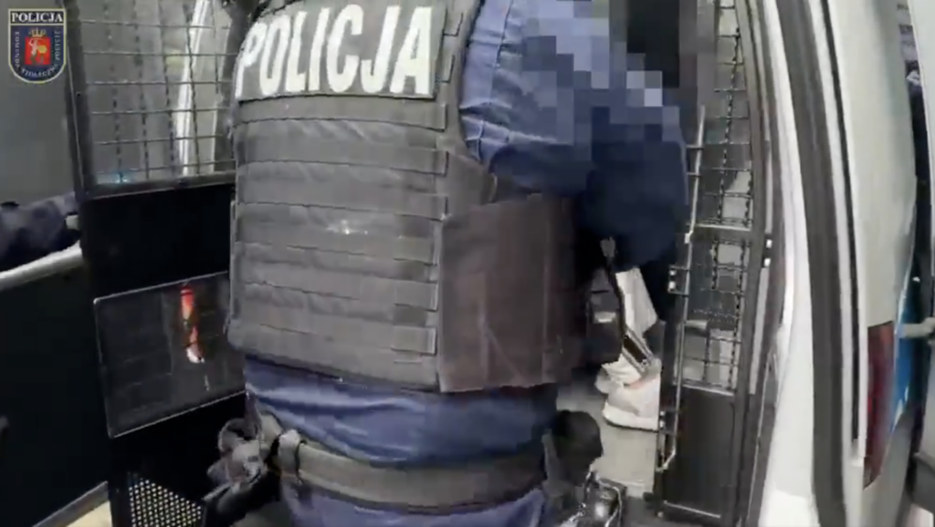 Policjant w kamizelce z napisem „POLICJA” prowadzi osobę z zamazaną twarzą do radiowozu.
