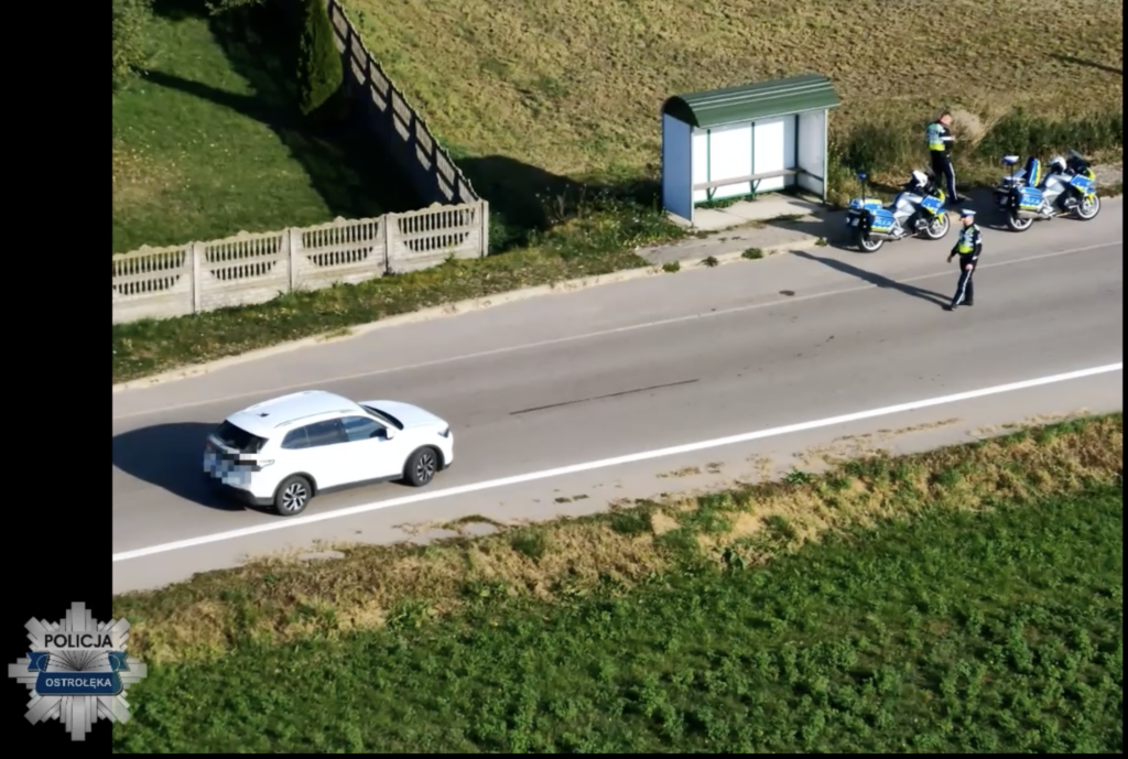 Biały samochód mija policjanta stojącego na drodze przy radiowozach motocyklistów i przystanku autobusowym.