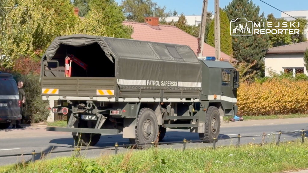 Wojskowa ciężarówka z napisem „PATROL SAPERSKI” jedzie ulicą wśród domów i drzew.