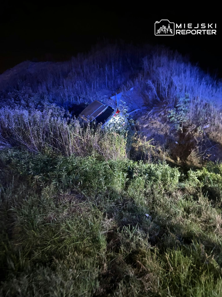 Samochód osobowy leży w zarośniętym rowie, oświetlony niebieskim światłem nocą.