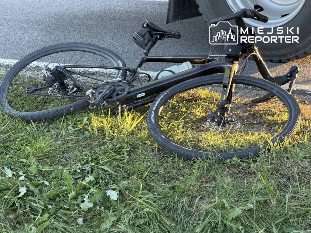 Czarny rower leży na trawie obok koła samochodu stojącego na asfaltowej drodze.