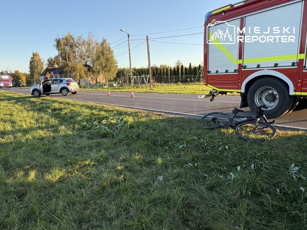 Rower leży przy wozie strażackim na poboczu drogi, obok zatrzymanego samochodu i pachołków drogowych.