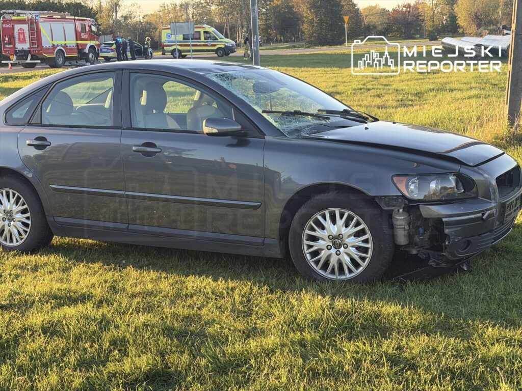 Uszkodzony szary samochód osobowy stoi na trawie, a w tle pracują strażacy i ratownicy medyczni.