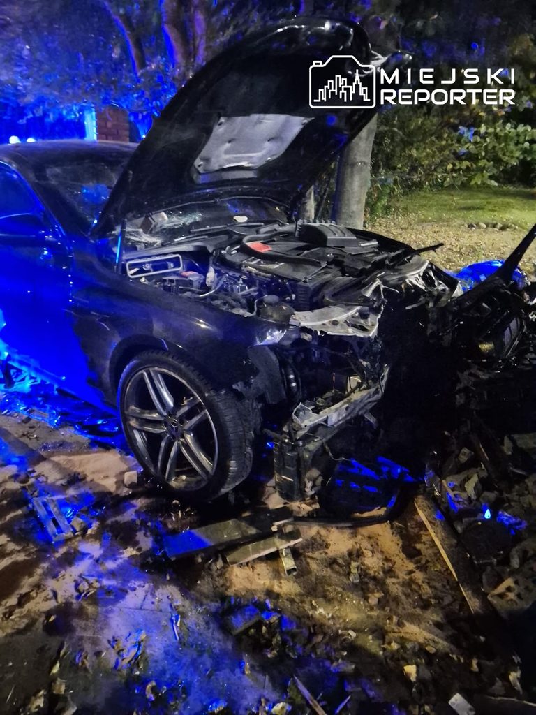 Rozbity czarny samochód z otwartą maską i poważnie uszkodzonym przodem, oświetlony niebieskimi światłami nocą.