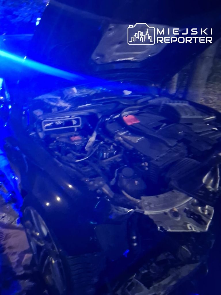 Uszkodzony samochód z otwartą maską, oświetlony niebieskimi światłami służb podczas nocnego wypadku