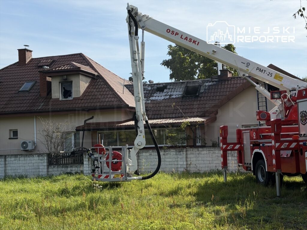 Strażacki podnośnik przy domu z uszkodzonym dachem i okopconym oknem po pożarze.