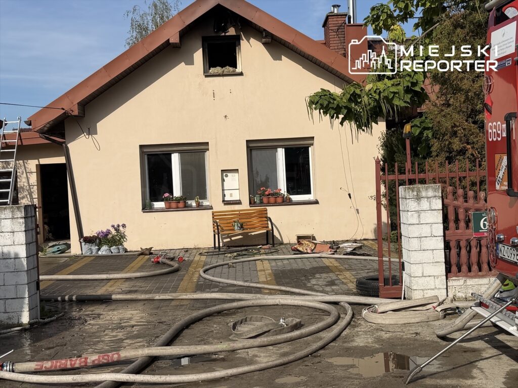 Węże strażackie przed domem z uszkodzonym oknem na poddaszu po pożarze, obok widoczny wóz strażacki.
