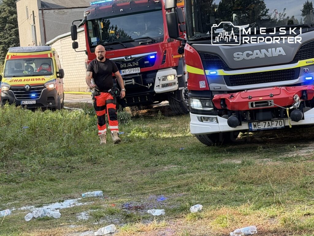 Strażak w odblaskowych spodniach stoi przed wozami strażackimi na trawiastym terenie obok budynku.