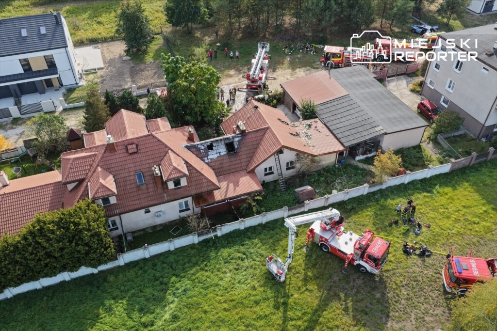 Strażacy prowadzą działania przy domu z uszkodzonym dachem, otoczonym wozami strażackimi i drabinami pożarniczymi.